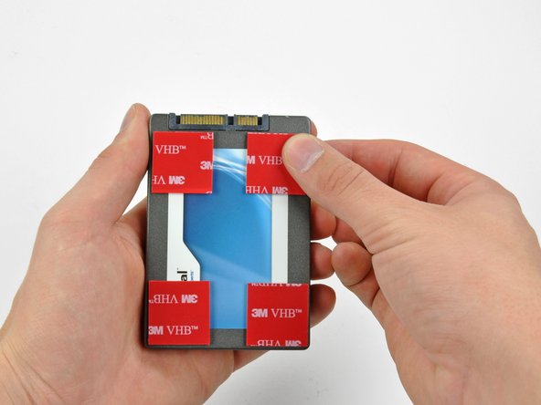 Image 3/3: Peel the backing off one side of the double-sided tape and place the pieces at the four corners of the hard drive.