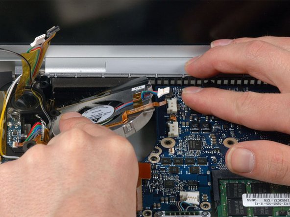 Image 1/1: Hold the logic board down with one hand and use your other hand to lift the left fan up from its housing. There is a piece of black tape securing the fan to the heat sink. Carefully peel this tape up from the heat sink as you lift the fan up.